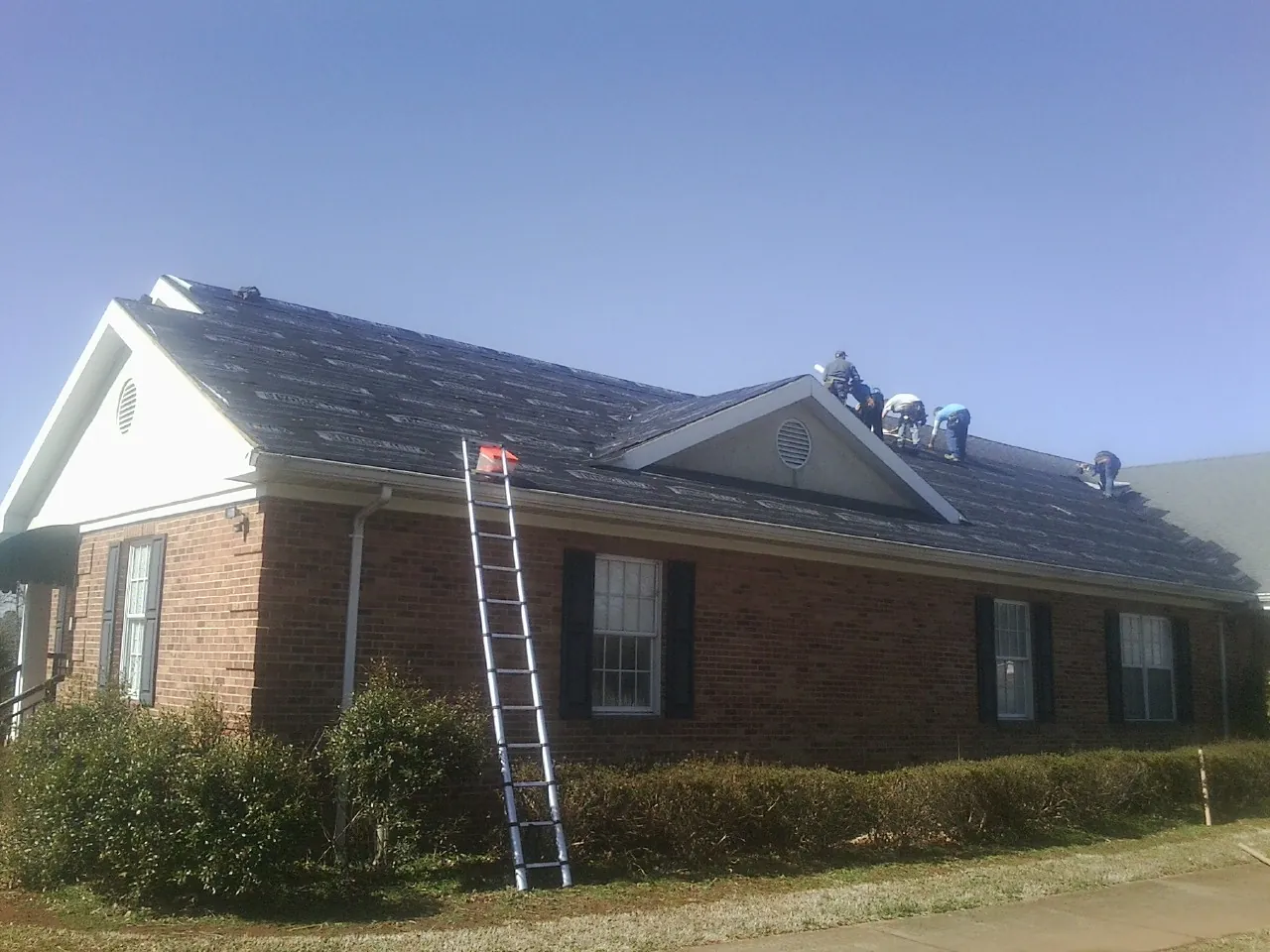 Crew tearing shingles off a brick ranch roof with a ladder set — Metal Roof Repair work by our crew in Colville