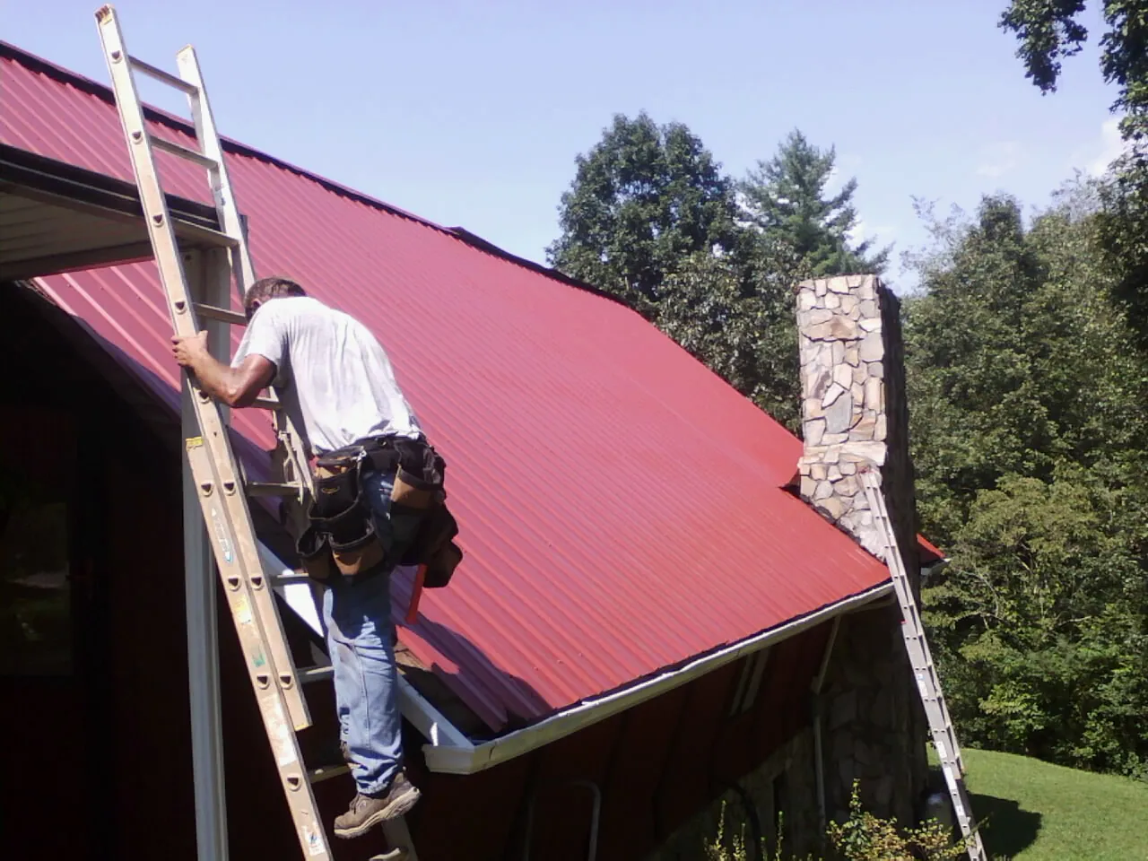 Roofer on a ladder beside a red metal roof and stone chimney — Soffit & Fascia Repair by Roofing Brothers in Colville